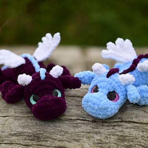 May include: Two plush dragon toys made of soft yarn. One dragon is deep purple with light blue accents and teal eyes, while the other is light blue with dark purple accents and pink eyes. Both feature white crocheted wings and small horns.