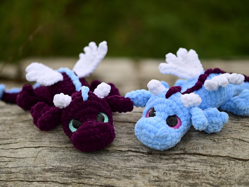 May include: Two plush dragon toys made of soft yarn. One dragon is deep purple with light blue accents and teal eyes, while the other is light blue with dark purple accents and pink eyes. Both feature white crocheted wings and small horns.