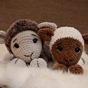 May include: Two crocheted stuffed sheep toys, one brown and one white, lying on a white fluffy surface. The sheep have black eyes and brown noses.