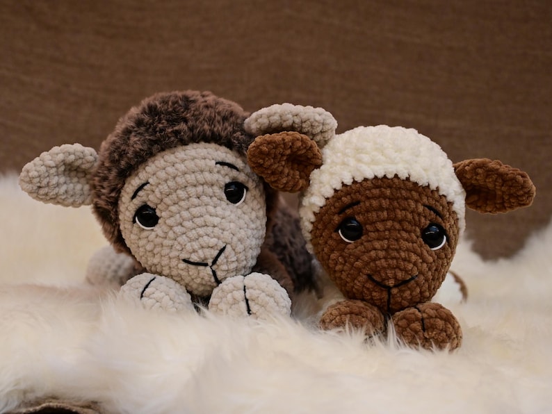 May include: Two crocheted stuffed sheep toys, one brown and one white, lying on a white fluffy surface. The sheep have black eyes and brown noses.