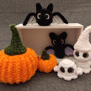May include: A collection of crocheted Halloween decorations, including a pumpkin, two bats, and two ghosts. The pumpkin is orange with a green stem, the bats are black with purple accents, and the ghosts are white with black eyes.