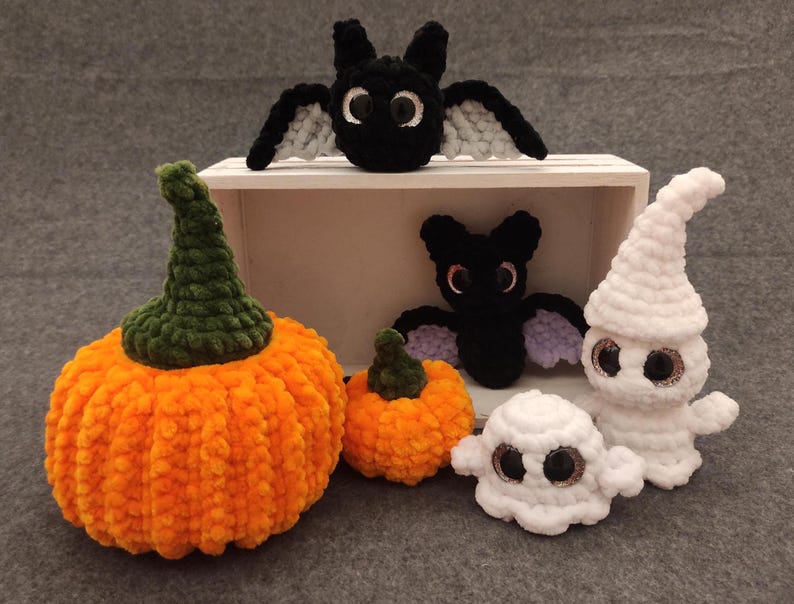 May include: A collection of crocheted Halloween decorations, including a pumpkin, two bats, and two ghosts. The pumpkin is orange with a green stem, the bats are black with purple accents, and the ghosts are white with black eyes.