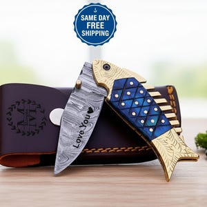 Op de afbeelding: Een decoratief vouwmes met een visvormig handvat. Het handvat is goud en blauw en het lemmet heeft een uniek patroon met de woorden "Love You". Achter het mes ligt een bruine leren etui met initialen.