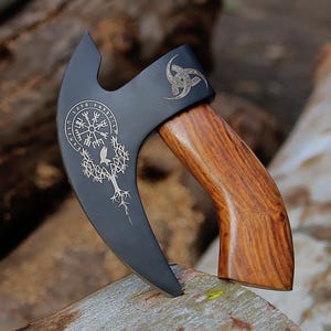 May include: A black axe with a wooden handle. The axe head features intricate engraved designs, including a compass rose, a tree, and a symbol. The handle is made of polished wood. The axe is displayed on a wooden surface.