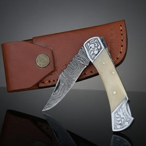 May include: A Damascus steel folding knife with a cream-colored handle and decorative silver accents. The knife is partially open, revealing the patterned blade. It rests beside a brown leather sheath with a decorative metal button.