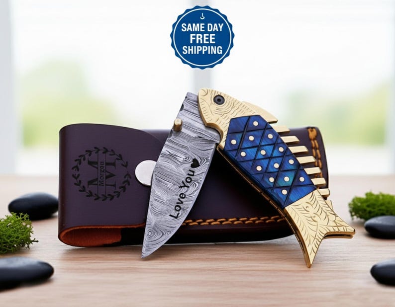 Op de afbeelding: Een decoratief vouwmes met een visvormig handvat. Het handvat is goud en blauw en het lemmet heeft een uniek patroon met de woorden "Love You". Achter het mes ligt een bruine leren etui met initialen.