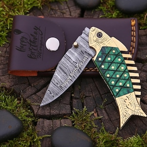 Op de afbeelding: Een decoratief vouwmes met een visvormig handvat. Het lemmet heeft een opvallend patroon en de tekst "Happy Birthday Jass". Het handvat is groen en goudkleurig, samen met een bruin lederen etui met de tekst "Happy Birthday Morgen".