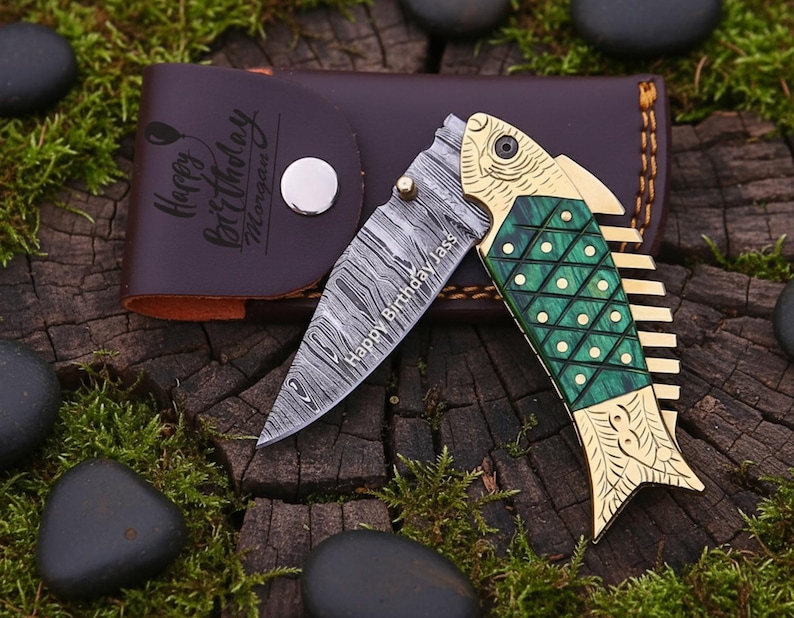 Op de afbeelding: Een decoratief vouwmes met een visvormig handvat. Het lemmet heeft een opvallend patroon en de tekst "Happy Birthday Jass". Het handvat is groen en goudkleurig, samen met een bruin lederen etui met de tekst "Happy Birthday Morgen".