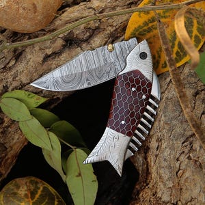 Op de afbeelding: Een vouwmes met een visvormig handvat en een patroon op het lemmet. Het handvat heeft een rood en bruin honingraatontwerp. Het lemmet is zilverkleurig met een uniek patroon. Het mes ligt op een natuurlijke houten achtergrond met bladeren.