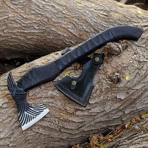 May include: A black handled axe with a silver blade and a black leather sheath. The axe has a decorative pattern on the blade and handle.