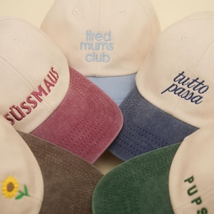 Peut inclure: Plusieurs casquettes de baseball de couleurs crème, bleu et bordeaux sont présentées. Chaque casquette comporte du texte brodé dans différentes polices et langues, dont "tired mums club" et "tutto passa". Les casquettes ont des visières incurvées.