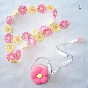 May include: A pink and yellow daisy chain lace trim and a pink flower-shaped measuring tape. The measuring tape is white with black markings and extends to 50 cm.
