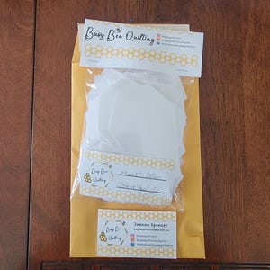 May include: A yellow envelope with a white label that says "Busy Bee Quilting" and a bee logo. Inside the envelope is a package of white hexagon fabric pieces. A business card with the text "Joanne Spencer" and the same bee logo is also visible.