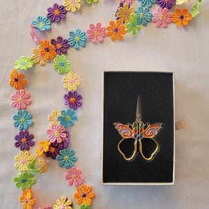 May include: A colourful floral trim with a butterfly-shaped pair of scissors in a black box. The box has a white label with the number #1 printed on it.