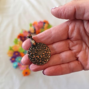 May include: A circular, bronze pendant featuring a floral pattern. The pendant has a small, clear gem at the top and is held in a hand. The background is blurred, with colourful flowers.