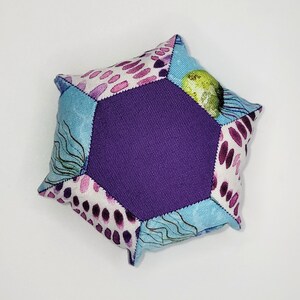 May include: A hexagonal fabric cushion with a central purple panel. The edges are made of patterned fabric in blue, white, and purple. The cushion has a patchwork design with abstract patterns and shapes, showcasing a handcrafted aesthetic.