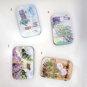 May include: Four rectangular metal tins with floral and landscape designs. The tins feature images of lavender fields, a bicycle, a bottle, and roses. The tins are in various colours, including purple, green, and brown.