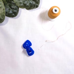 May include: A top-down view shows sewing supplies on a white surface. A spool of orange thread, a needle, and two blue thimbles are visible. Dark green leaves are in the upper left corner. The thimbles have a dotted pattern.