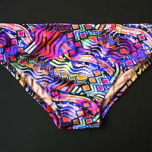 May include: Vibrant, colorful swim briefs with a bold, abstract pattern. The design features a mix of geometric shapes and wavy lines in shades of pink, purple, orange, blue, and green. The briefs have a light tan interior lining and a black trim.