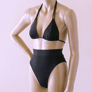Puede incluir: Conjunto de bikini negro sobre un maniquí. La parte superior es de estilo triangular con finas tiras halter. La parte inferior de cintura alta ofrece cobertura total. El traje de baño está hecho de tela negra lisa.