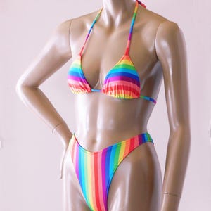 May include: A rainbow-striped bikini set. The top is a triangle style with thin straps, and the bottoms are high-cut. The fabric features vertical stripes in all the colors of the rainbow, creating a vibrant and eye-catching look.