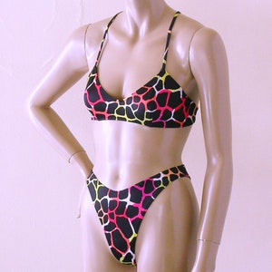 Op de afbeelding: Zwarte tweedelige bikini met een patroon van gele, roze en witte onregelmatige vormen. De top heeft een halternek ontwerp en de broek is hoog uitgesneden. De set wordt getoond op een mannequin.