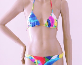 May include: A brightly coloured bikini set with an abstract design. The triangle top and low-rise bottoms feature blue, red, yellow, and purple brushstrokes. The halter straps and edges are trimmed in coordinating colours, creating a bold and eye-catching swimwear piece.