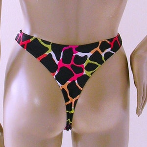 May include: Black thong-style swimsuit bottom with a colorful abstract pattern. The pattern features shapes in pink, orange, green, and white against a black background. The swimsuit is displayed on a mannequin.