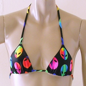Puede incluir: Top de bikini negro con estampado repetido de caras de alienígenas coloridas. Las caras de los alienígenas son en tonos de rojo, rosa, amarillo, verde y azul. Las correas y los bordes del top también están decorados con acentos coloridos.