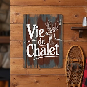 May include: A rustic metal sign with the words "Vie de Chalet" and a deer illustration. The sign is mounted on a wooden wall, alongside a snowshoe, a red and black plaid blanket, and a mug.
