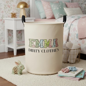 Peut inclure: Un panier à linge crème avec des poignées noires. Le panier porte le nom "EMMA" en lettres colorées à motifs au-dessus des mots "DIRTY CLOTHES" en noir. Une figurine de lapin décorative et des livres sont à proximité.