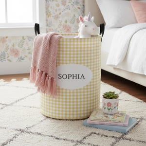 May include: A yellow and white gingham patterned laundry hamper with black handles. The hamper contains a pink throw blanket with fringe and a stuffed unicorn. The name "SOPHIA" is printed on the front. A small potted plant and books sit nearby.