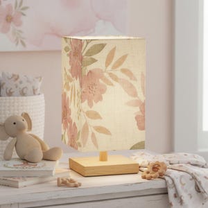 May include: A rectangular table lamp with a floral pattern in pink and beige. The lamp has a wooden base and a linen shade. The lamp is on a white surface, with a stuffed animal and books nearby. The lamp is designed for a nursery.