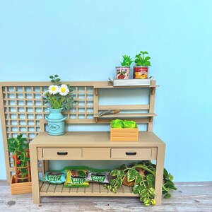 May include: A miniature wooden garden workbench with two drawers, a shelf, and a trellis. The workbench has three bags of Happy Gro Co. seeds, a small potted plant, and two cans of tomatoes on the shelf. The workbench is on a light brown wooden surface.