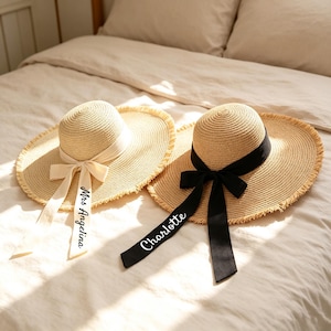 May include: Two straw sun hats with wide, frayed brims. One hat features a cream-coloured ribbon tied in a bow with the text Mrs Angelina printed on the tail. The other hat has a black ribbon tied in a bow with the text Charlotte printed on the tail.
