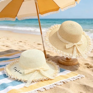 May include: Two straw sun hats with cream-colored lace trim and bows sit on a beach towel. One hat has the name "Sophia" printed on it. A yellow and white striped beach umbrella provides shade on the sandy beach.