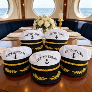 Op de afbeelding: Vijf wit-zwarte kapiteinspetten met gouden gevlochten rand en gele geborduurde laurierbladeren op de klep. Elke pet is voorzien van een zwart ankericoon en de tekst CAPTAIN gevolgd door een naam: OWEN, DANIEL, HENRY, ETHAN of LIAM.