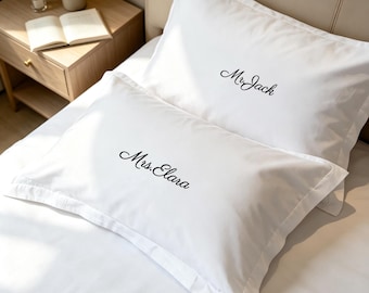 May include: Two white rectangular pillowcases featuring embroidered black script text. The top pillowcase displays the name Mr. Jack, while the bottom pillowcase features the name Mrs. Elara. Both pillowcases have a clean, smooth fabric finish.