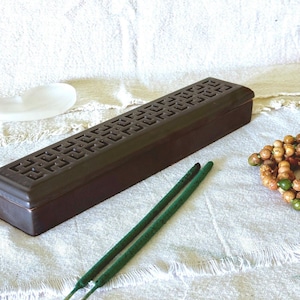 May include: A brown rectangular incense burner with a patterned top, two green incense sticks, and a beaded necklace on a white cloth. A white, shell-shaped dish is also present.