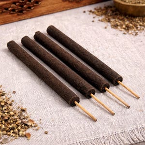 May include: Four dark brown incense sticks with wooden stems are arranged diagonally on a textured, off-white surface. The incense sticks are cylindrical and have a rough texture. Dried herbs and spices are scattered around the sticks.