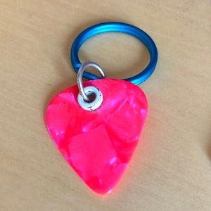 May include: A bright pink guitar pick attached to a blue metal key ring. The pick is on a wooden guitar.