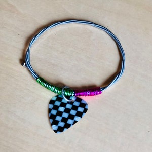 May include: A necklace made from a guitar string with a black and white checkered guitar pick pendant. The string is wrapped into a circular shape, with green and pink accents near the pendant.