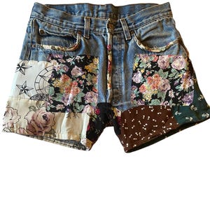 May include: Blue denim shorts with patchwork floral and patterned fabric accents. The shorts feature a classic five-pocket design with a button and zipper closure. The patchwork includes floral, star, and dot patterns.