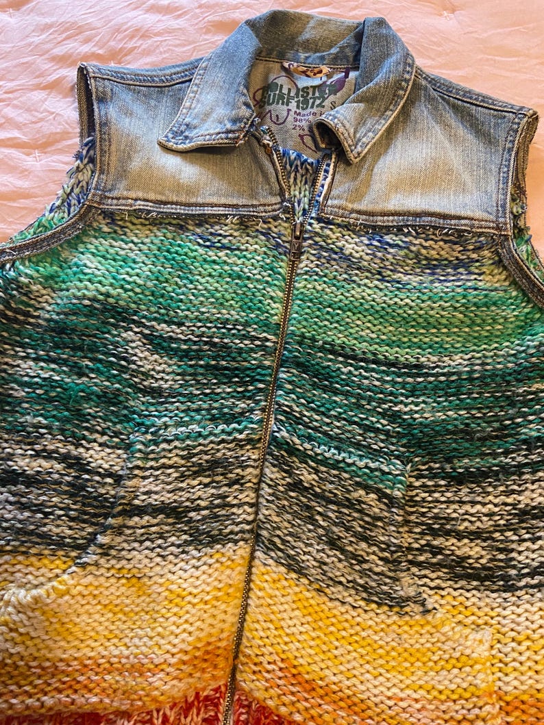 May include: A sleeveless denim vest with a knitted front. The vest features a light blue denim collar and shoulders, transitioning to a vibrant, striped knit in shades of green, yellow, and red. A full-length zipper runs down the front.