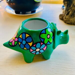 May include: A green ceramic triceratops planter with colorful flower designs. The planter has a wide opening for plants and is painted with pink, blue, yellow, and green flowers. The dinosaur planter is a unique decorative item.