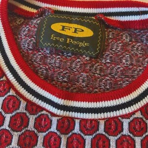 May include: Close-up of a red, white, and black sweater with a repeating pattern of red smiley faces. The sweater has a ribbed neckline with red, white, and black stripes. A tag reads "FP free People".