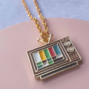 May include: Gold chain necklace with a vintage television pendant. The television is brown with a gold frame and has a colourful test pattern on the screen.