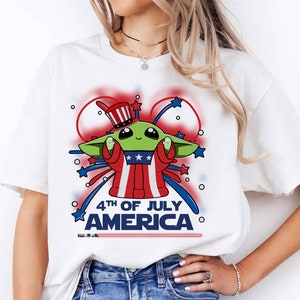 Puede incluir: Camiseta blanca con un personaje extraterrestre verde que lleva un sombrero rojo, blanco y azul y sostiene un fuego artificial. El texto "4th of July America" est&aacute; impreso debajo del personaje.