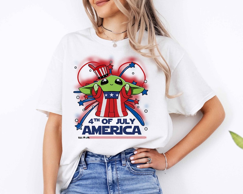 Puede incluir: Camiseta blanca con un personaje extraterrestre verde que lleva un sombrero rojo, blanco y azul y sostiene un fuego artificial. El texto "4th of July America" est&aacute; impreso debajo del personaje.