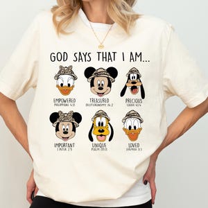 A white t-shirt with a graphic design featuring six cartoon faces of Disney characters, along with text that says "God says that I am..." and Bible verse references.